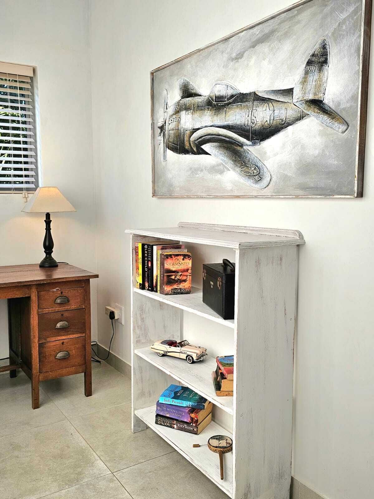A room with a desk, a lamp, a bookshelf with books, and a framed airplane painting.