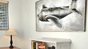 Ein Zimmer mit Schreibtisch, Lampe, Bücherregal mit Büchern und einem Bild von einem Flugzeug im Bilderrahmen.