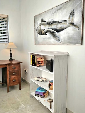 A room with a desk, a lamp, a bookshelf with books, and a framed airplane painting.