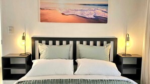 Ein Bett mit weißer Bettwäsche, zwei Nachttischlampen, ein Kopfteil und ein Bild einer Strandlandschaft darüber.