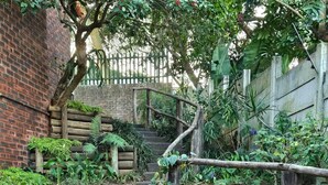 Eine steinerne Treppe führt zu einem hölzernen Zaun, umgeben von üppiger Vegetation und einer Backsteinmauer.