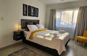 A modern bedroom with a bed, bedside tables, a nightstand, a window with curtains, and artwork on the wall.