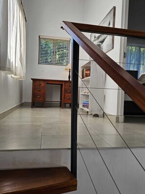 A room with a staircase, a desk, a window with blinds, and a bookshelf.