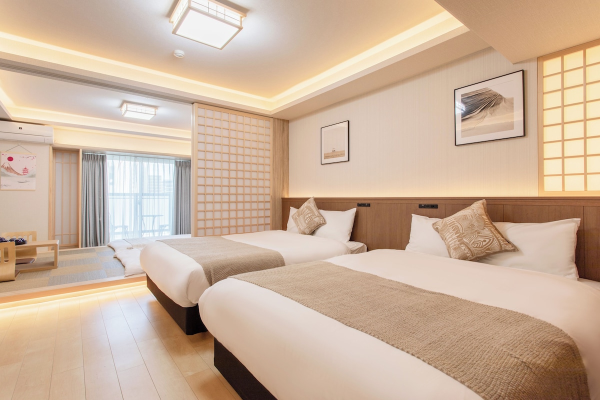Ein Hotelzimmer mit zwei Betten, einem Futon, einem Shoji-Bildschirm und einem traditionellen japanischen Kunstwerk.