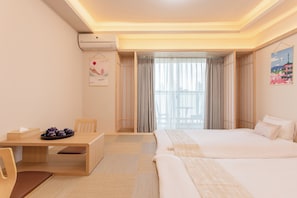 A hotel room with a bed, a desk, and a chair. There is a window with curtains, a wall-mounted air conditioner, and a painting.