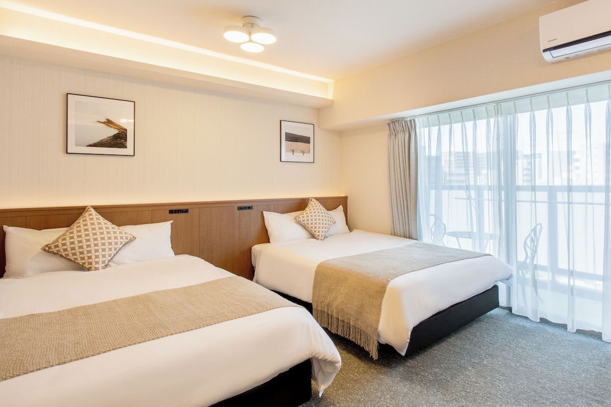 Ein Hotelzimmer mit zwei Betten, einem Holz-Kopfteil, einem großen Fenster mit durchscheinenden Vorhängen und einer Wand-Klimaanlage.