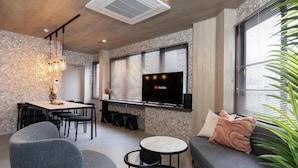 Een moderne woonkamer met een eethoek, een flatscreen televisie aan de muur en een groot raam met jaloezieën.