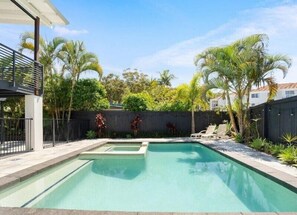 Ein rechteckiger Swimmingpool mit einer kleinen Whirlpool-Ecke, umgeben von einem Holzzaun, Palmen und einem überdachten Patio.