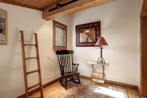 A room with a wooden ladder, a rocking chair, a small table with a lamp, a mirror, and a window with a view of trees. - Historic Downtown Grand Lake Cabin with Hot Tub Walk to Beach and Shops (Grand Lake)