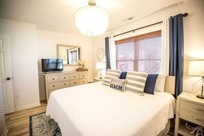 Une chambre à coucher comprenant un lit, une commode, un téléviseur, une fenêtre avec des rideaux et un lustre.