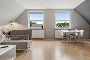 A modern loft bedroom with a sloped ceiling, a grey sofa, a dining table with chairs, and large windows. - Holiday apartment in Nexø close to beach and port (Nexø)