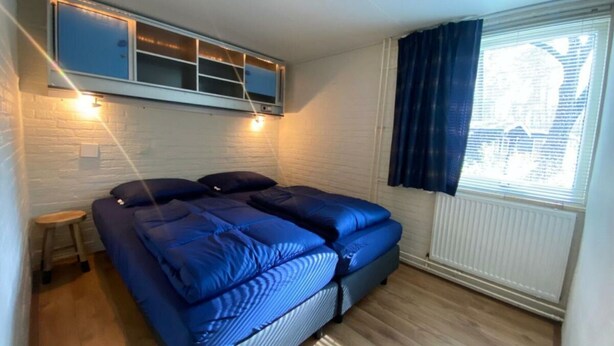 A bedroom with a bed, a small stool, a window with curtains, and a wall-mounted shelf. - Holiday Home in Beekbergen With Terrace (Beekbergen)