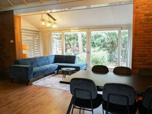 A modern room with a blue sectional sofa, a dining table with chairs, and large sliding glass doors leading to a garden. - Holiday Home in Beekbergen With Terrace (Beekbergen)