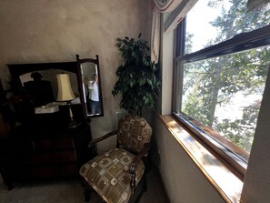 Ein Zimmer mit Stuhl, Schminktisch, Spiegel, Lampe und Fenster mit Blick auf Bäume.