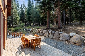 Un patio con una mesa y sillas de madera, rodeado por un muro de piedra y rocas grandes, con un área boscosa al fondo.