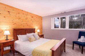 A bedroom with a wooden wall, a bed with white bedding, two bedside lamps, a window, and a chair with folded towels.