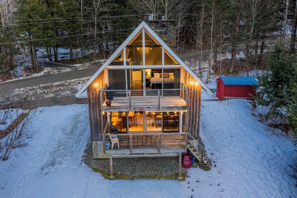 A two-story cabin with a balcony, a stone roof, and a red shed in the background.