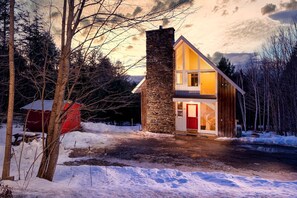 Une maison à deux étages, dotée d’une cheminée en pierre, d’une porte rouge et d’un escalier apparent menant à l’étage supérieur.