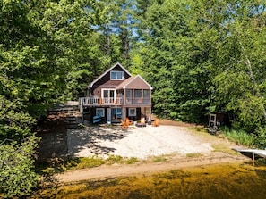A two-story cabin with a balcony, situated on a sandy beach surrounded by dense trees.