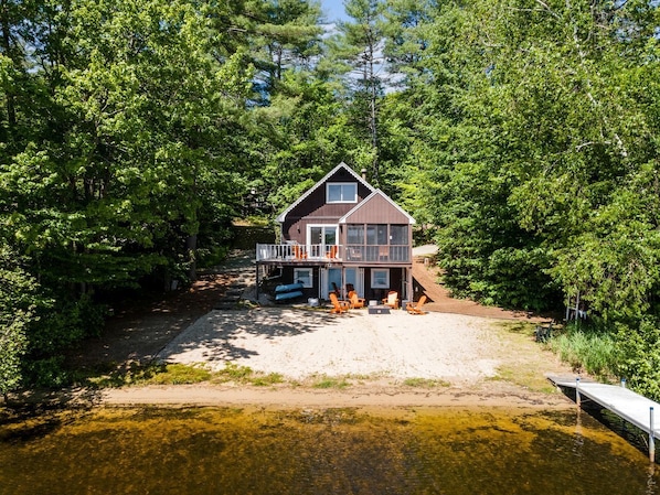 A two-story cabin with a balcony, situated by a lake surrounded by dense trees.