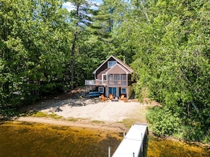 A cabin with a deck, surrounded by dense forest, near a body of water.