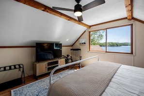 Un dormitorio con cama, televisor, ventilador de techo, ventana con vista a un lago y vigas de madera.