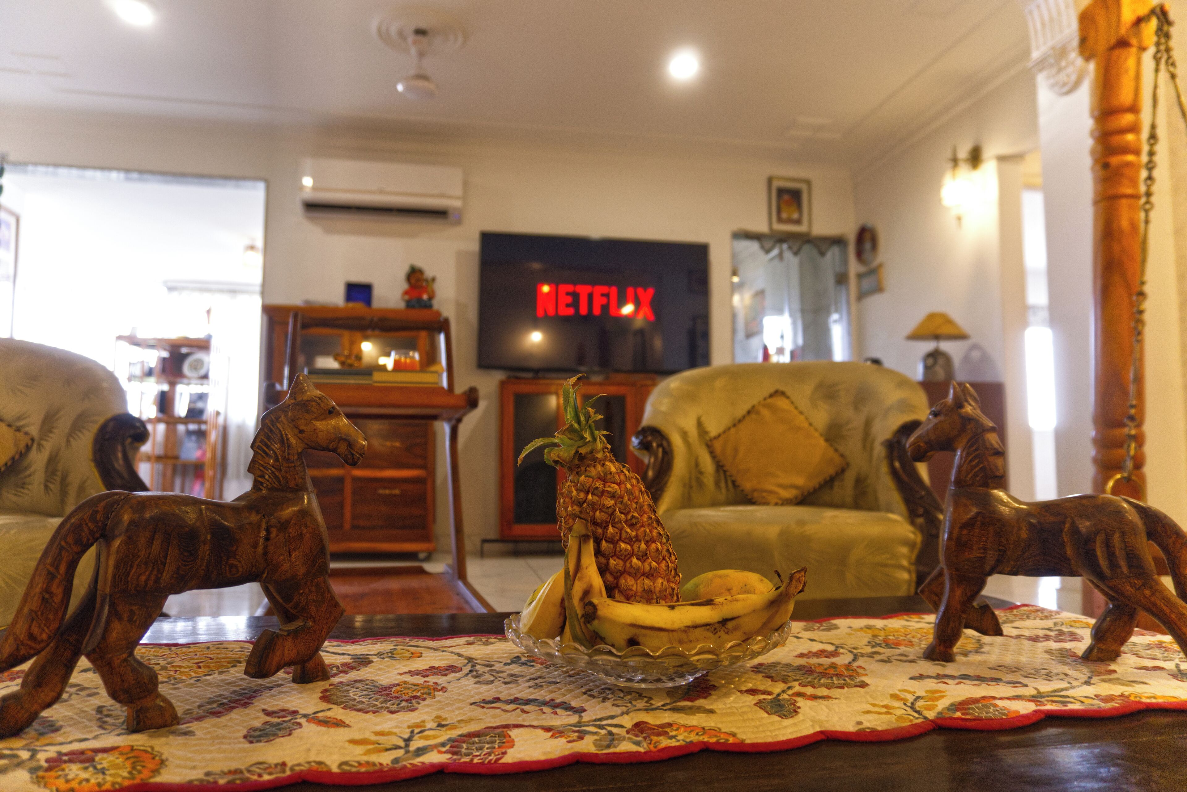 A living room with a television displaying the Netflix logo, a wooden horse figurine, a pineapple, and a banana on a table.
