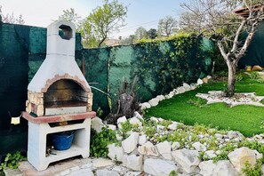 Ein gemauerter Außenofen mit Backsteinunterbau, umgeben von einer Steinmauer und Grünpflanzen.