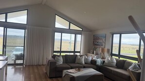 A spacious living room with a large sectional sofa, a coffee table, and a view of the countryside through large windows. - Meadow Lees - Linen, Wi-Fi, Pet Friendly (Venus Bay)