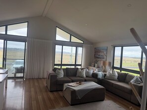 A spacious living room with a large sectional sofa, a coffee table, and a view of the countryside through large windows. - Meadow Lees - Linen, Wi-Fi, Pet Friendly (Venus Bay)