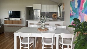 A modern kitchen with white cabinets, a stainless steel refrigerator, and a dining area with a white table and chairs. - Meadow Lees - Linen, Wi-Fi, Pet Friendly (Venus Bay)