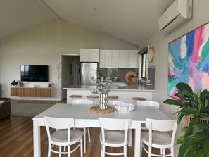 A modern kitchen with white cabinets, a stainless steel refrigerator, and a dining area with a white table and chairs. - Meadow Lees - Linen, Wi-Fi, Pet Friendly (Venus Bay)