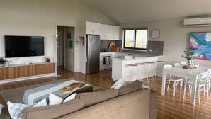 A modern living room with a flat-screen TV, a sofa, and a dining area with a white table and chairs. - Meadow Lees - Linen, Wi-Fi, Pet Friendly (Venus Bay)