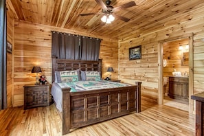 A wooden cabin bedroom with a bed, nightstands, a ceiling fan, and a bathroom visible through an open door. - Secluded Rustic Cottage in Gatlinburg, Tennesee - Ideal Spot for a Family Vacation in the Great Smoky Mountains National Park (Gatlinburg)
