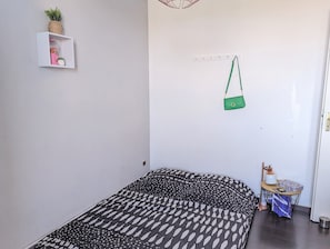 Ein Schlafzimmer mit einem Bett, einem kleinen Regal mit einer Pflanze, einer hängenden Tasche und einem Tisch mit einer Lampe.