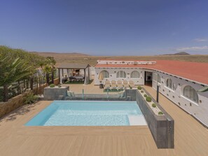 Ein Poolbereich mit Blick auf eine Wüstenlandschaft, ein überdachter Außenbereich zum Sitzen und ein Gebäude mit Bogenfenstern.