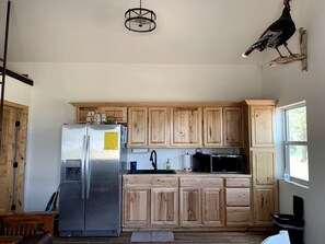 A kitchen with wooden cabinets, a stainless steel refrigerator, a microwave, and a hanging bird. - Charming, Glamping Cabin with Barbecue Grill, Perfect for Wildlife Watching in Raton, New Mexico (Raton)