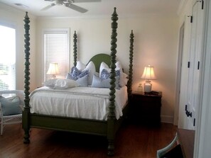 Uma cama com dossel e estrutura verde, roupa de cama branca e almofadas listradas em azul e branco. Há uma mesa de cabeceira com uma luminária e uma cadeira com assento branco.