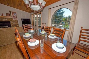 A dining room with a wooden table set for four, surrounded by wooden chairs, a stone fireplace, and a large window with a view of trees.