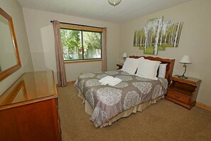 A bedroom with a large bed, a nightstand with a lamp, a dresser, and a window with curtains.