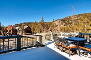 A snow-covered deck with outdoor furniture, a view of a mountainous landscape, and buildings in the background.