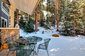 A covered outdoor seating area with a stone fireplace, surrounded by snow-covered ground and tall pine trees.