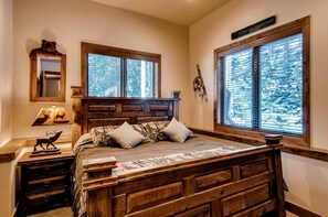 A bedroom with a wooden bed, two windows with blinds, a mirror, a lamp, and a sign above the windows.