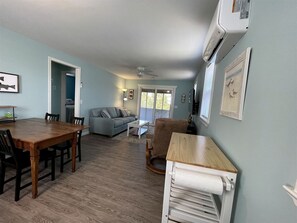 Ein geräumiges Wohnzimmer mit einem Holzessttisch, einer Couch und einem Schaukelstuhl. Es gibt einen weißen Schrank mit Handtuchspender und einen Deckenventilator.