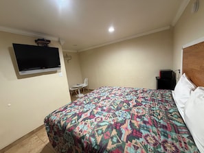 A bedroom with a bed, a television mounted on the wall, a small table with chairs, and a nightstand with a lamp.
