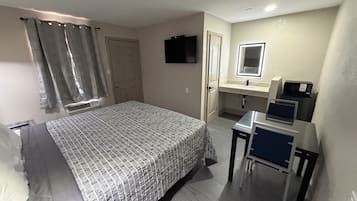 Un dormitorio con cama, escritorio, silla, televisor y espejo, además de un baño con lavamanos e inodoro.