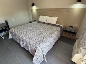 A neatly arranged hotel room with a large bed, a desk, a chair, a bedside table, and a wall-mounted lamp.