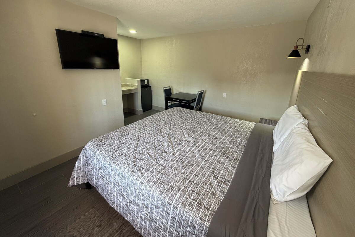 A hotel room with a bed, a desk, a chair, a TV, and a small kitchenette.