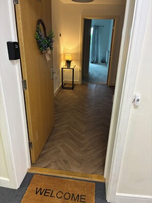 Ein Korridor mit Holzboden, einer Fußmatte, einem kleinen Tisch mit Lampe und einer Tür mit Schloss.