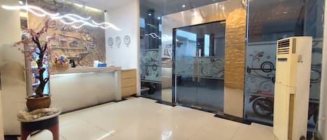 A hotel reception area with a reception desk, a potted plant, a wall clock, and a large air conditioning unit.